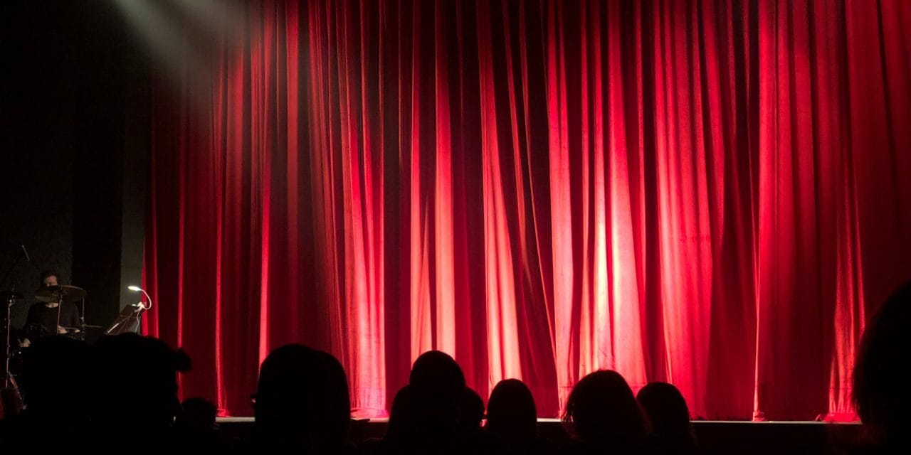 A red curtain occupies the stage blocking view of the performance as silhouettes of an audience are visible in the foreground with a drummer in a dimly lit venue.