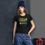 A young woman stands against a dark wall wearing a black t-shirt with the text "HAVE A LUCKY ST. PATRICK'S DAY!" and a blue beanie  accompanied by industrial elements nearby.