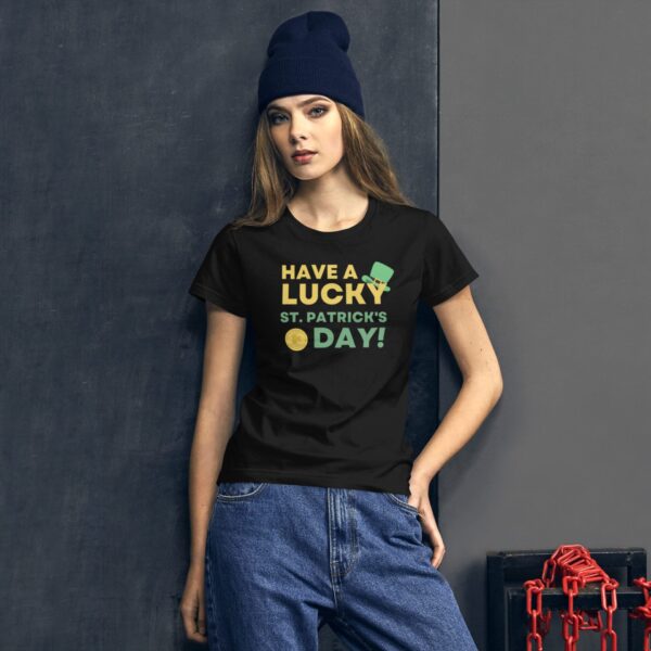 A young woman stands against a dark wall wearing a black t-shirt with the text "HAVE A LUCKY ST. PATRICK'S DAY!" and a blue beanie  accompanied by industrial elements nearby.