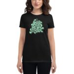 A woman wearing a black t-shirt displays the phrase "Enjoy Every Moment" in stylized green lettering  standing in a neutral background  highlighting the shirt's message and design.