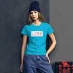 A model stands confidently wearing a bright turquoise t-shirt with the word LOVE prominently displayed in red. She is accessorized with a dark blue beanie in a modern urban setting.