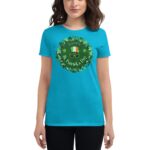 A woman wears a bright blue t-shirt featuring a circular design with shamrocks and a festive green hat with glasses that says HAPPY St. Patrick's Day  surrounded by greenery.