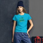 A young woman stands confidently wearing a bright blue t-shirt with the text "TOO CUTE TO PINCH" and an arrow design  paired with denim jeans against a dark background.