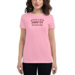 A woman wears a pink t-shirt featuring the text "LOVE IS MY favorite ADVENTURE" in black. She stands in a neutral setting  smiling gently.