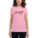 A woman wears a pink t-shirt featuring the phrase "BELIEVE IN LOVE" in bold black letters with decorative elements  standing against a neutral background  smiling gently.