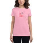 A woman wears a pink t-shirt featuring a graphic of two hands forming a heart shape above the word love in red cursive  standing against a neutral background.
