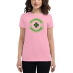 A woman wears a pink t-shirt featuring a green clover design with the text celebrating St. Patrick's Day and themes of love and luck amidst a plain background.