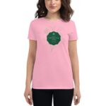 A woman wears a pink t-shirt featuring a green label with the text Life's like a cup of tea surrounded by simple line-drawn botanical illustrations  standing against a neutral background.