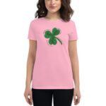 A woman wears a pink t-shirt featuring a green four-leaf clover design with the text "Prosperity in business Luck in life Love in relationships" surrounding it against a neutral background.