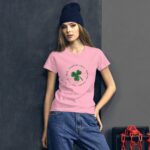 A young woman stands confidently wearing a pink t-shirt with a green four-leaf clover graphic surrounded by words on a dark wall with a chain in the background.