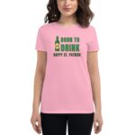 A woman stands wearing a pink t-shirt featuring the text "BORN TO DRINK HAPPY ST. PATRICK" alongside an illustration of a beer bottle and glass  celebrating St. Patrick's Day.