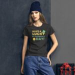 A woman stands confidently wearing a dark t-shirt with the text "HAVE A LUCKY ST. PATRICK'S DAY!" along with a green hat and golden coin graphic against a gray wall and a red chain backdrop.
