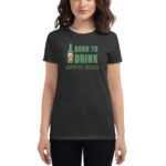 A woman is wearing a dark gray t-shirt featuring the text "BORN TO DRINK HAPPY ST. PATRICK" with an illustration of a beer bottle in the center.