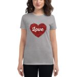 A woman stands wearing a light gray t-shirt featuring a red heart design with the word Love in white at the center  conveying a positive emotional message in a casual setting.