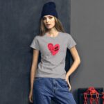 A young woman wears a gray t-shirt featuring a red heart graphic with the text "SELF LOVE" and an illustration. She poses against a dark wall  with a beanie on her head.