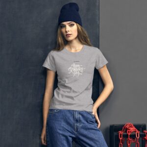 A woman stands confidently wearing a gray t-shirt with a playful design that says Happy St Patrick's Day. She has long hair and a navy hat  against a dark background with chains.