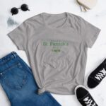A gray t-shirt with the text Happy St. Patrick's Day in green is displayed alongside blue jeans and black sneakers on a light surface  with round sunglasses nearby.
