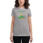 A woman is wearing a gray t-shirt featuring a colorful cartoon character celebrating St Patrick's Day surrounded by festive illustrations like shamrocks and confetti in a casual indoor setting.