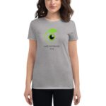 A woman wears a gray t-shirt featuring a green logo with a black circle in the center and text "SAINT PATRICKS 2024" below it. She stands against a neutral background.