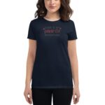A woman stands wearing a dark navy t-shirt featuring the text "LOVE IS MY favorite ADVENTURE" in a playful font  against a neutral backdrop emphasizing the shirt's design.