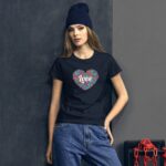 A young woman stands against a textured dark wall wearing a black t-shirt with a colorful heart design stating love and loose blue jeans with a dark beanie on her head.