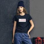 A young woman in a black t-shirt with "LOVE" printed on it stands confidently against a dark wall. She wears a dark beanie and blue jeans  conveying a casual yet expressive style.