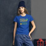 A young woman stands against a dark wall wearing a blue t-shirt with a festive message for St. Patrick's Day and casual jeans a dark beanie adorns her head
