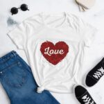 A white t-shirt featuring a red heart design with the word Love inside is displayed alongside blue jeans and black sneakers on a light surface with sunglasses nearby.