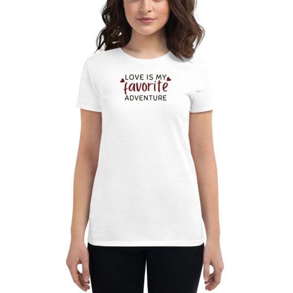 A woman is wearing a white t-shirt featuring the text "LOVE IS MY favorite ADVENTURE" in a playful font  standing in a neutral environment with no distinct background details.