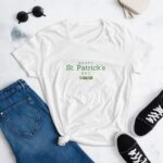 A white t-shirt featuring the text "HAPPY St. Patrick's DAY" in green is styled with knotted at the waist next to blue jeans and black sneakers on a light surface with sunglasses and a small bag.