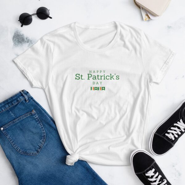 A white t-shirt featuring the text "HAPPY St. Patrick's DAY" in green is styled with knotted at the waist next to blue jeans and black sneakers on a light surface with sunglasses and a small bag.