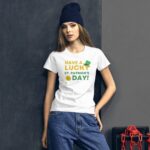 A woman stands wearing a white t-shirt with a festive message while posing against a textured gray wall. She is accessorized with a dark beanie and denim jeans. The shirt reads HAVE A LUCKY ST. PATRICK'S DAY!