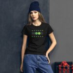 A young woman stands against a textured dark wall wearing a black t-shirt with green graphics that read "MOOD" surrounded by small leaf designs paired with blue jeans and a knitted beanie.