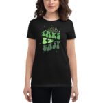A woman is wearing a black t-shirt featuring the phrase TAKE IT EASY in large green  playful lettering along with two cartoonish characters and stars in a casual setting.