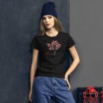 A young woman wearing a black T-shirt with a tree design made of pink hearts stands against a textured gray background with a casual pose and a blue beanie on her head.