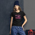 A woman stands against a dark textured wall wearing a black t-shirt with the pink text "WINE IS MY VALENTINE" and denim jeans while a beanie rests on her head.