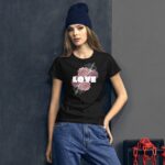 A young woman stands against a textured gray wall wearing a black t-shirt with a floral design and the word love in bold letters  paired with loose blue jeans and a dark beanie.