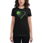 A woman wears a black t-shirt featuring a green heart and shamrock design with the text Happy St Patrick's Day  celebrating the holiday in a casual setting.