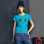 A model wearing a bright turquoise t-shirt with the word "MOOD" and graphic emojis is posed against a dark grey wall. She has long hair and wears a dark beanie.