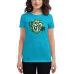 A woman stands wearing a bright turquoise t-shirt displaying the word "EXPECTATION" in bold black letters surrounded by a circular green leaf design against a neutral background.