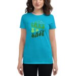 A woman stands wearing a bright turquoise t-shirt with the phrase "TAKE IT EASY" in a playful green font surrounded by whimsical illustrations and small stars in a casual setting.