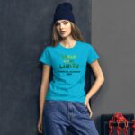 A woman poses confidently wearing a bright turquoise t-shirt with the text "DREAM WITHOUT LIMITS HAPPY ST. PATRICKS 2024" alongside casual blue jeans against a dark gray wall with chains nearby.