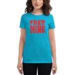 A woman stands wearing a bright blue t-shirt featuring bold red text that reads "BE MINE" surrounded by red heart shapes in a neutral background.