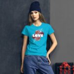 A young woman stands confidently against a dark wall wearing a bright turquoise t-shirt with the word LOVE surrounded by floral designs and dressed in denim jeans with a beanie.