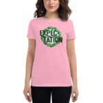 A woman in a pink t-shirt stands  showcasing a circular graphic with the word "EXPECTATION" in bold black letters surrounded by green clovers  set against a neutral background.