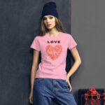 A young woman stands confidently wearing a pink t-shirt with a heart design and the word "LOVE" on it in black. She has long hair and a navy beanie. The background is dark and minimalistic with hints of red chains.