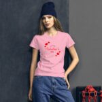 A young woman stands against a dark textured backdrop wearing a pink t-shirt that reads Happy Valentine’s Day surrounded by red hearts while dressed in casual denim and a navy beanie.