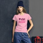 A young woman stands confidently wearing a pink t-shirt with the text "WINE IS MY VALENTINE" beside a dark wall  styled with casual jeans and a beanie  conveying a relaxed vibe.