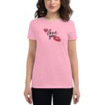 A woman stands wearing a light pink t-shirt featuring the words love you in black script accompanied by two red lips and hearts  set against a neutral background.
