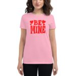 A woman wears a light pink t-shirt with bold red text that reads BE MINE surrounded by small heart shapes conveying a playful and romantic theme.